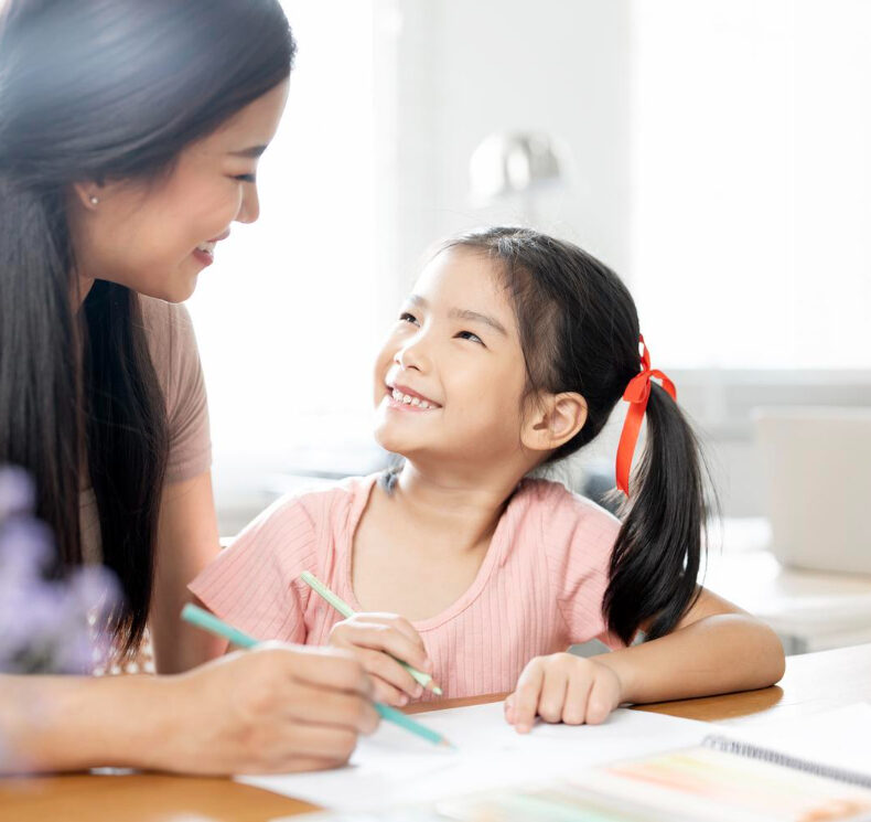 Girl with a red ribbon in her hair smiles at a woman as they draw together at a table with pencils and paper nearby.