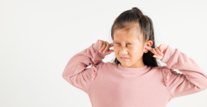 Young girl in a pink sweater covering her ears, eyes squeezed shut, showing discomfort or sensitivity to noise.