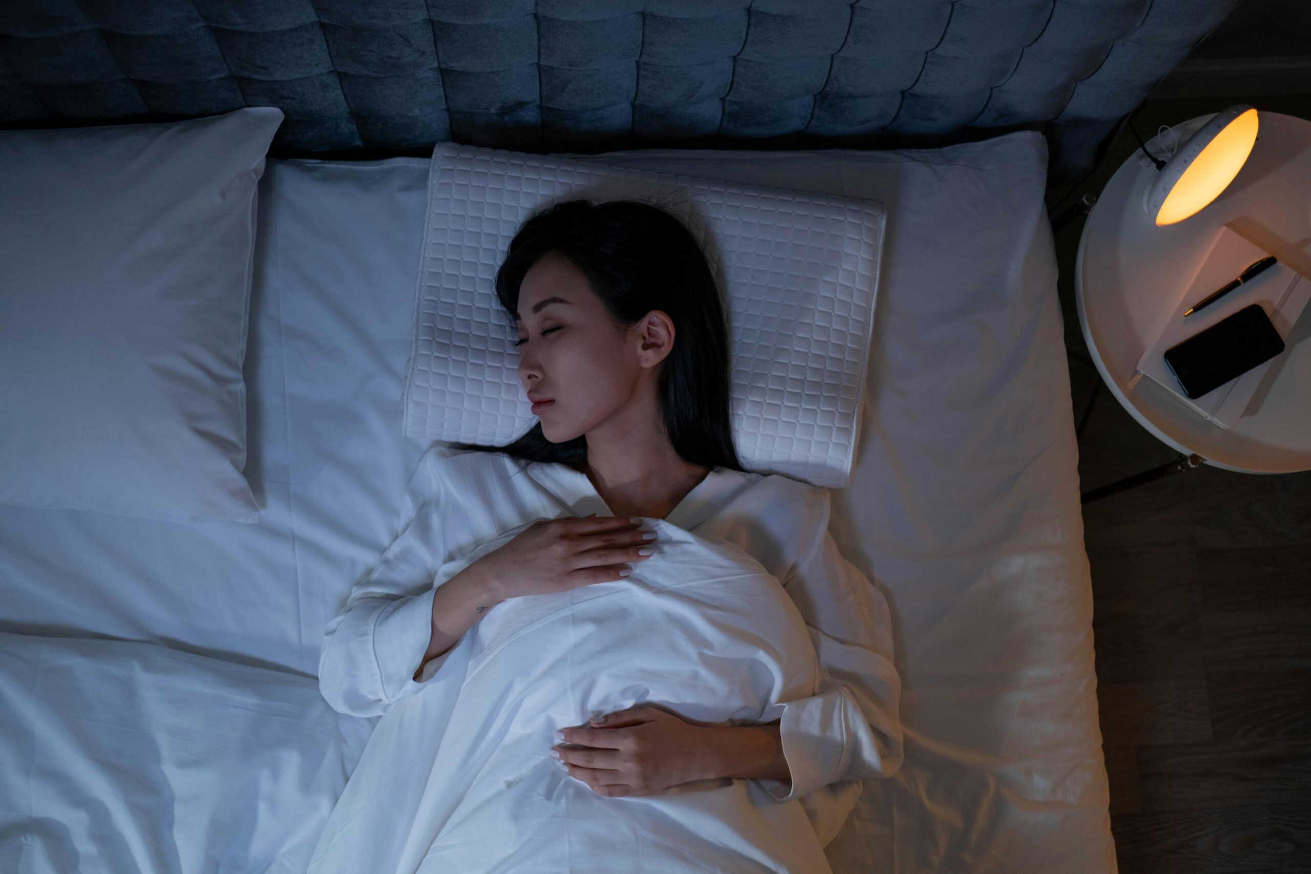 A person sleeping on a white-bedded bed, hands resting on chest, near a lit bedside lamp and a table with a phone and pen nearby