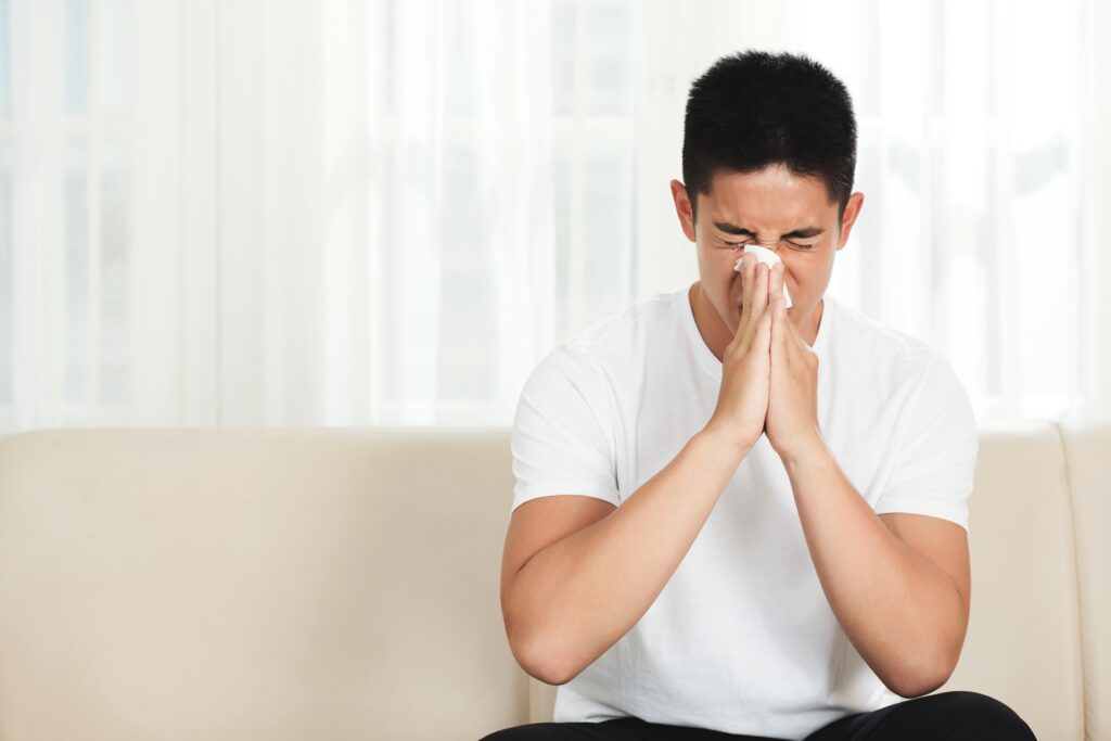 Young man sneezing or blowing his nose with tissue while sitting on a sofa indoors.