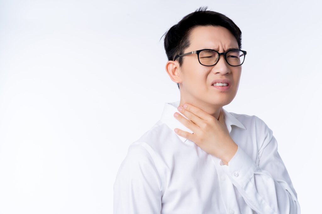 Man wearing glasses grimaces in discomfort while touching his throat with his right hand against a plain white background.
