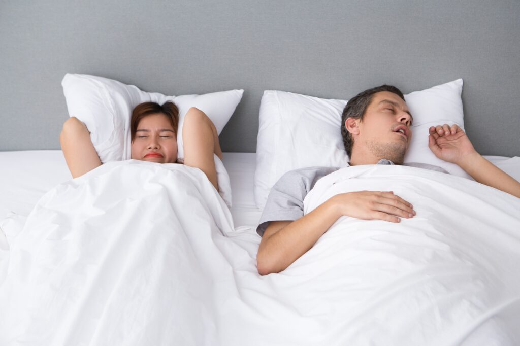 Couple sleeping in bed with white sheets and pillows, woman on the left and man on the right against a gray headboard.